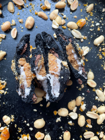 The "Dark Chocolate, Peanut Butter, Marshmallow" dessert from She means Biscuits features a blackened exterior with a creamy filling, nestled on a dark surface amid scattered peanuts, toffee bits, and hints of dark chocolate.