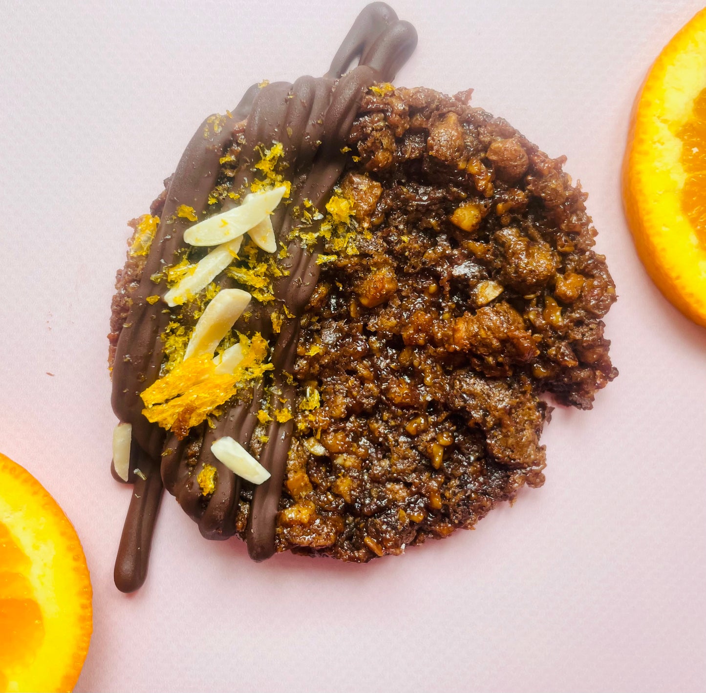 A close-up of a Salted Orange, Toasted Almond, Dark Chocolate (v+gf) cookie by She means Biscuits that is half covered with a dark chocolate drizzle, almond slivers, and orange zest. The cookie has a textured, crumbly appearance. Two orange slices frame the cookie on either side, adding a pop of color to the composition.