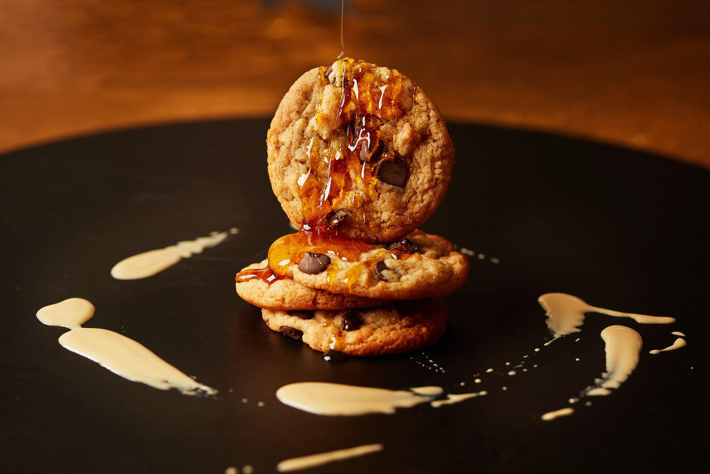 Three Kindness Cookies (Tahini, Maple, Choc Chunk) from She Means Biscuits are stacked on a black plate, drizzled with golden syrup. Artful swirls of tahini chocolate butter surround the cookies, adding to this delectable vegan treat. The background is softly blurred, highlighting the cookies as the main focus.