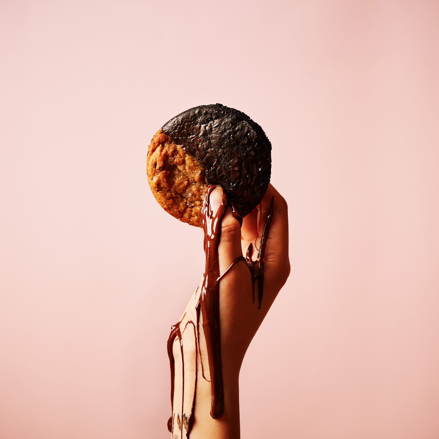 A hand holding a half-dipped Brookie, with melted chocolate cascading down the fingers, set against a light pink background, from She means Biscuits.