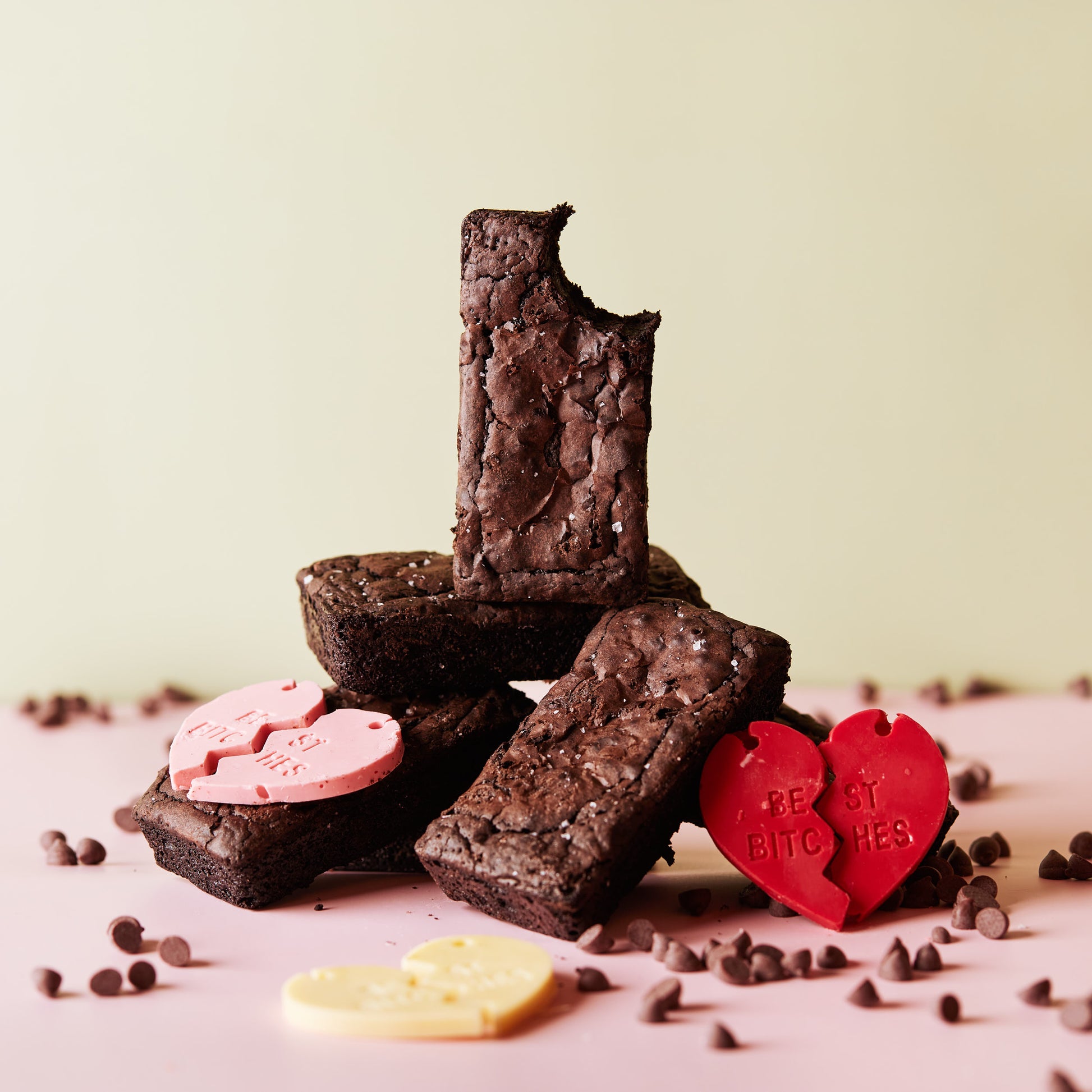 A stack of "She means Biscuits" Fudgy Brownies is arranged on a light pink surface, with the top one featuring a bite taken out. Two heart-shaped chocolates—one pink and one yellow—with sweet engravings are nestled among scattered dark chocolate chips.