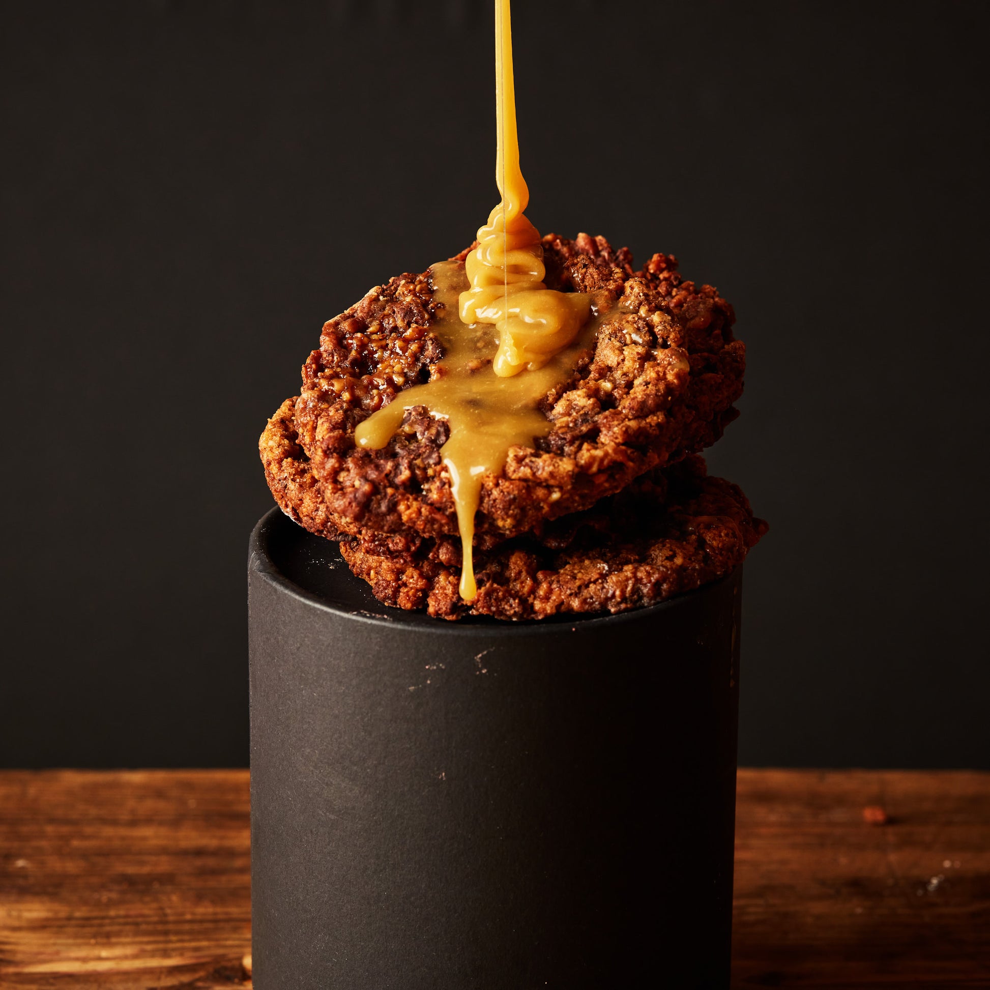 A stack of two Salty Cashews, Caramel, Choc Chunk (V&GF) cookies from She means Biscuits sits on a dark cylinder as creamy caramel is drizzled over them. The scene is set against a black background, with the wooden surface beneath adding a warm, rustic touch.