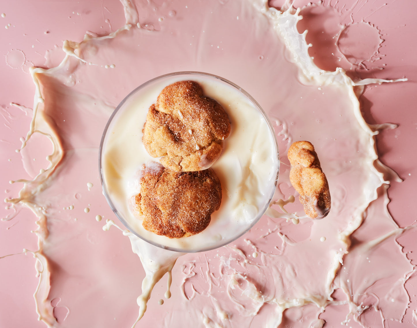 A bowl of milk with two Cinnamon Roll Biscuits from She means Biscuits rests on a pink surface. The milk splashes around the bowl, forming dynamic patterns across the background. A small, cloud-shaped piece of biscuit lies beside the bowl, enhancing this timeless treat.