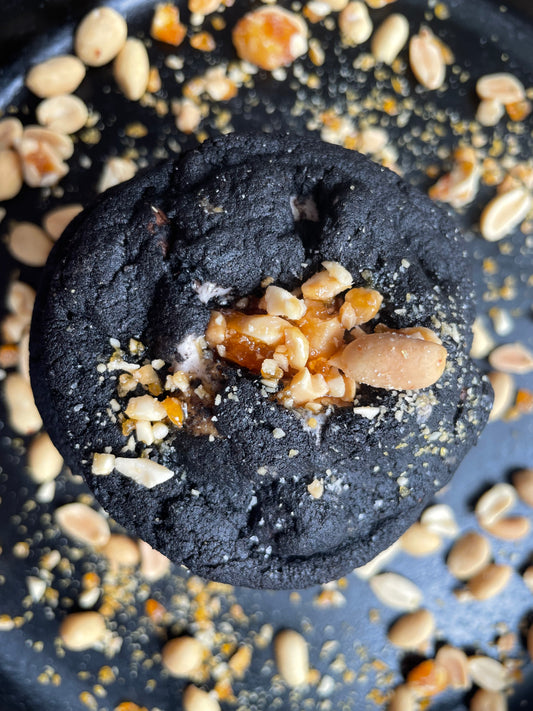 A close-up of the "Dark Chocolate, Peanut Butter, Marshmallow" treat by She means Biscuits, featuring a charred black muffin adorned with crushed peanuts, caramel bits, and flecks of dark chocolate. Whole peanuts are scattered around on the dark surface.