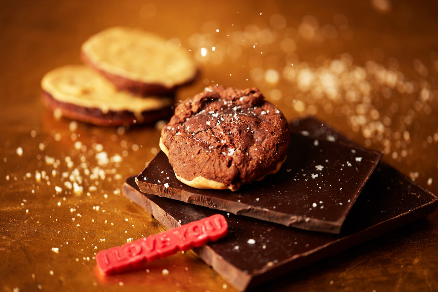 A cookie with chocolate and vanilla layers rests on a slab of dark chocolate, encircled by sprinkled sugar. Close by, you can find a Blonde Bottomed Brownie Biscuit (GF) by She means Biscuits, adorned with caramelized white chocolate. To the left sits a small red candy embossed with "I LOVE YOU," all enveloped in a warm, golden hue.