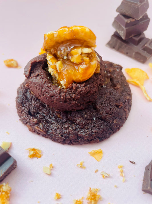 A Fancy Afghans (V+Gf) cookie from She means Biscuits, topped with caramelized nuts, sits on a light pink background. Surrounding it are chocolate chunks and small nut pieces, enhancing the delightful scene.