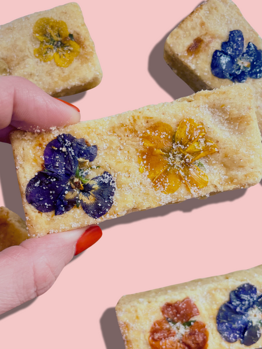 A hand with red nail polish holds a Buttery Shortbread cookie from She means Biscuits, adorned with colorful edible flowers. Other similar cookies are scattered on a pink background, each sprinkled with sugar.