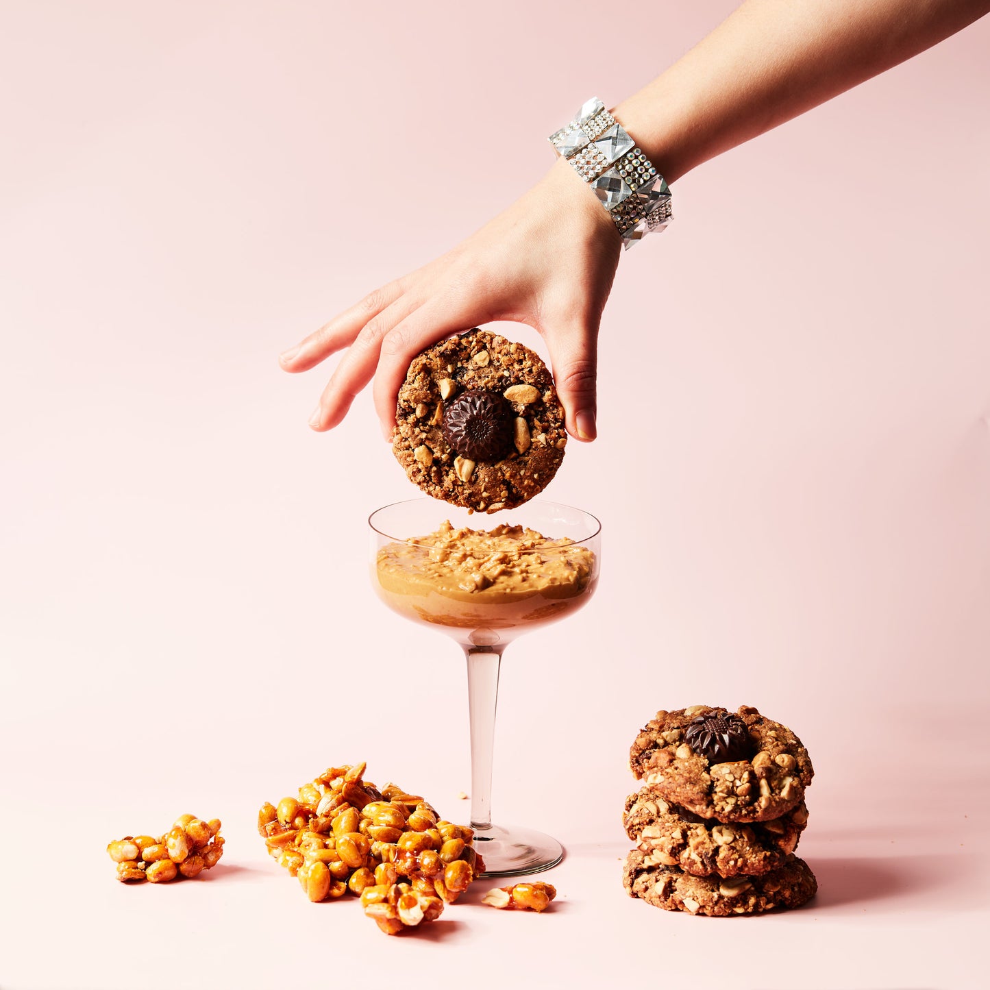 A hand adorned with a bracelet dips the Miso Peanut Butter (V&GF) vegan cookie from She Means Biscuits into a dessert-filled glass set against a light pink background. Beside the glass, there are caramel popcorn clusters and a stack of cookies featuring nuts and dark chocolate.