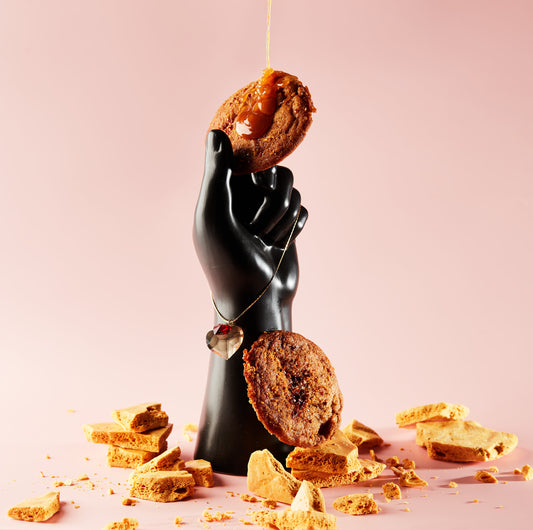 A black mannequin hand holds a biscuit from She means Biscuits' Honey Buttered Toast & Sea Salt line, dripping with caramel and sea salt. Another biscuit hangs elegantly from a necklace. Crumbled cookie pieces, infused with hints of Manuka honey, scatter across the pink surface.