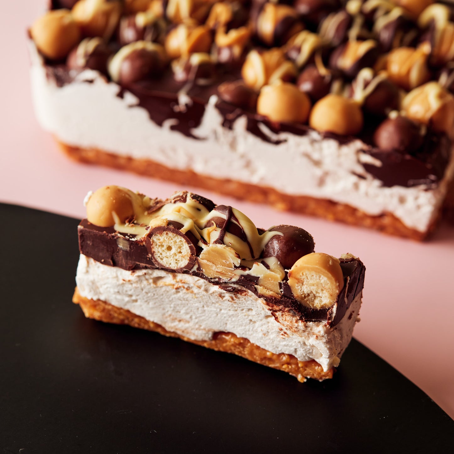 A close-up of a Marshmallow Crunch Slice by She means Biscuits on a black plate. It features a golden cornflake crunch base, topped with layers of white and dark chocolate, and is decorated with round chocolate sweets and nuts. In the background, there's a larger section of this gluten-free dessert.