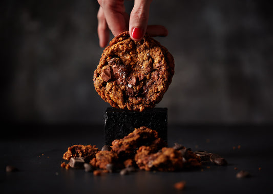 A hand with red nail polish holds a Hazelnut Choc (V&GF) cookie from She means Biscuits above a small black stone block. Crumbled pieces of the vegan and gluten-free cookie, along with rich chocolate chunks, are scattered on the dark surface below, creating an enticing treat.